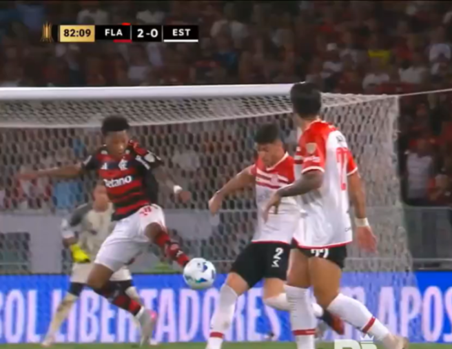 A soccer player in a red and black striped jersey kicking the ball towards the goal, with two opponents in white and red jerseys nearby.