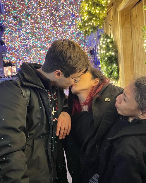 Two people kissing, and another person watching, against a backdrop of Christmas lights.