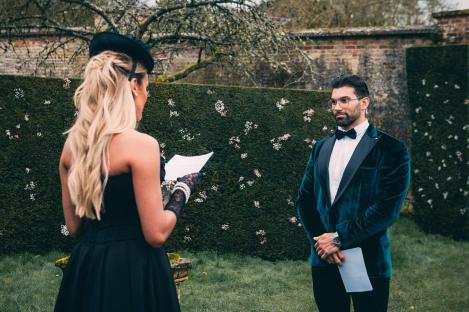 A woman in a black dress and hat reads from a piece of paper as a man in a teal velvet tuxedo looks on.