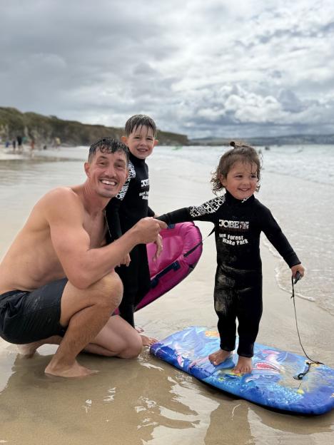 A father and his two young sons on a beach, one son standing on a boogie board.