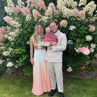 Family photo in front of hydrangeas; a woman in a peach ombre gown, a man in a beige suit, and their baby.