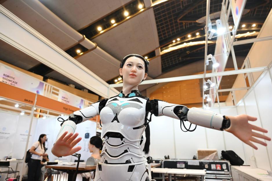 A humanoid robot with a human-like face and black hair tied back in a bun, wearing a white and black chest piece and white arm pieces, with wires visible on the joint of its left arm, standing with its arms outstretched in front of a booth at an electronics fair.