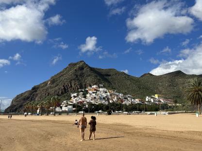 Couple walking on a sandy beach with a town on a hillside in the background.