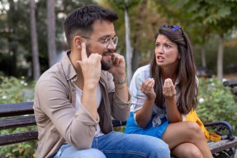 A woman debates with a man who covers his ears, highlighting tension and communication barriers, their differing expressions evident amidst a serene park backdrop.