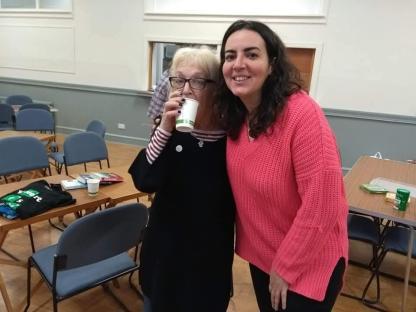 Two women standing in a room, one drinking from a cup.