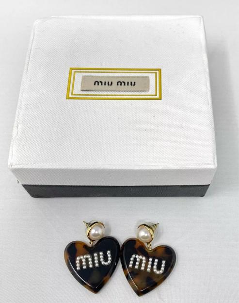 Pair of Miu Miu heart-shaped earrings made of tortoiseshell-patterned material, each with "Miu Miu" spelled out in pearls, next to a Miu Miu box.