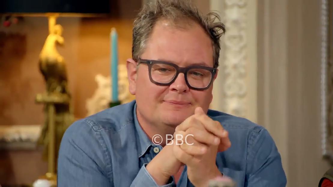 Alan Carr with dark-rimmed glasses and a denim shirt, with clasped hands.