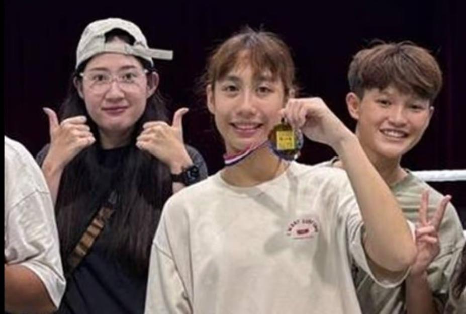 A female athlete holds up a medal while two others give thumbs up and peace signs.