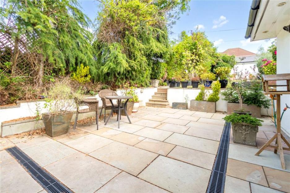 A paved patio area with a table and two chairs, surrounded by potted plants and greenery, leading up to a tiered garden with steps.