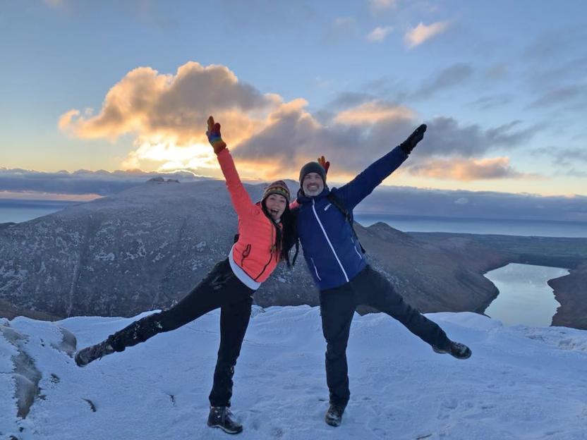 Couple joyfully posing on snowy mountaintop at sunset.