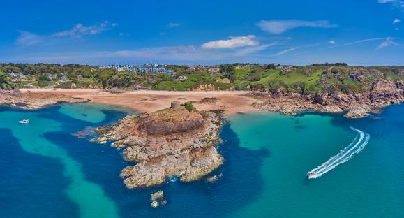 Aerial view of Portelet Bay, Jersey, with a sandy beach, turquoise water, and a small tower on a rocky outcrop.