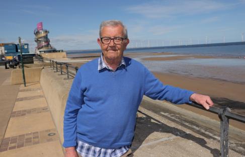 A man stands by a railing overlooking a beach with wind turbines in the background.
