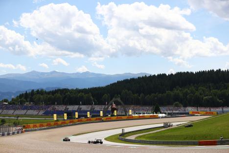Formula One cars racing on a track with spectators and mountains in the background.