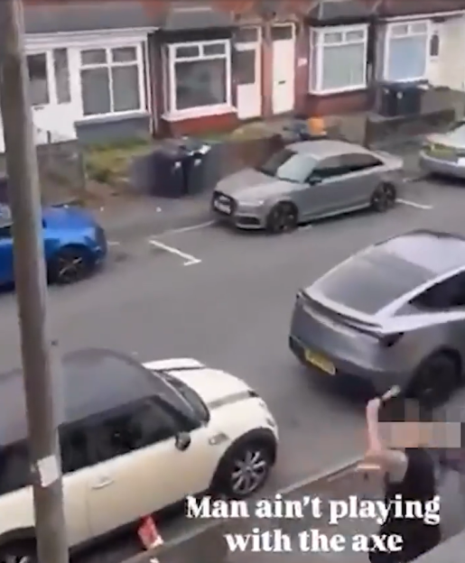 A person holding a blurred object, possibly an axe, standing near parked cars on a residential street.
