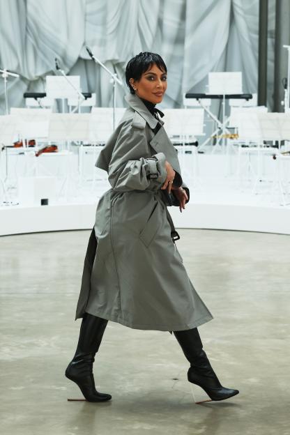 Kim Kardashian wearing a gray trench coat and black boots, looking over her shoulder at the Maison Margiela show.