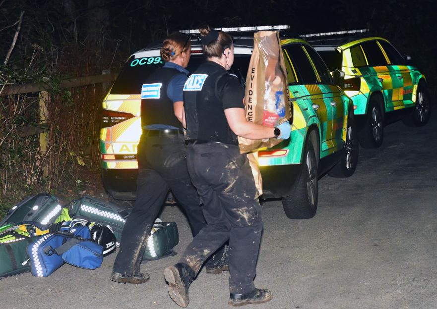 Police officers carrying evidence bag near emergency vehicles.
