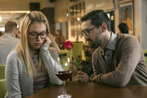 A man offering a red rose to a sad woman in a bar.
