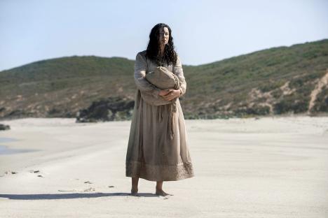 Woman in a long dress holding a bag on a beach.