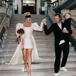 Couple and child descending a grand staircase; the woman is in a white dress and carries a bouquet.
