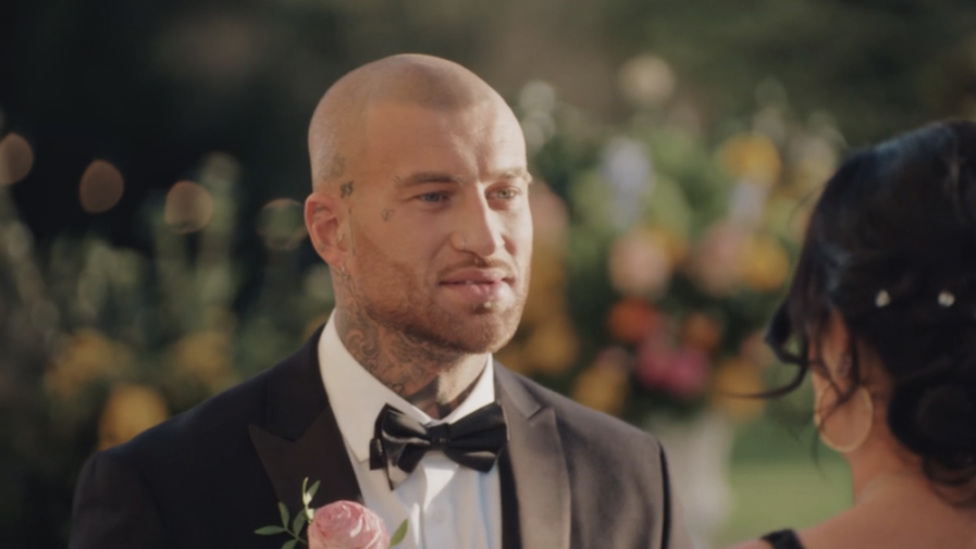 A man in a tuxedo with tattoos on his head and neck looks at a woman.