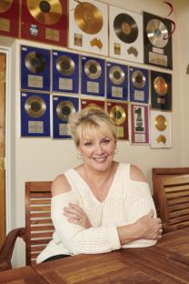 Cheryl Baker sitting at a table with many gold records on the wall behind her.