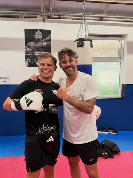 David Bentley in a boxing gym with a bloodied face, arm around a man in boxing gloves, in front of a boxing bag and a poster of Floyd Mayweather.