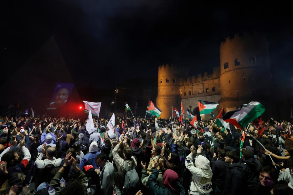 A large crowd of pro-Palestinian protestors raising their hands and waving Palestinian flags at night with an illuminated ancient Roman wall in the background.