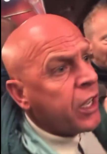 A bald man with blue eyes and a white turtleneck under a blue jacket yells.