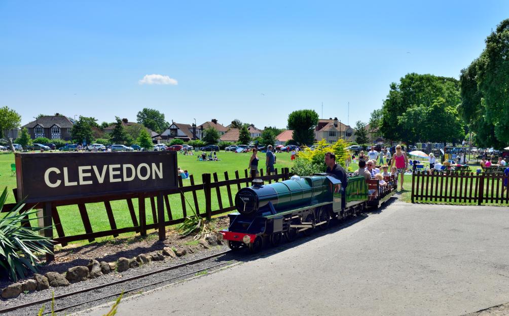 A green miniature train with passengers moving past a "CLEVEDON" sign in a park with families.