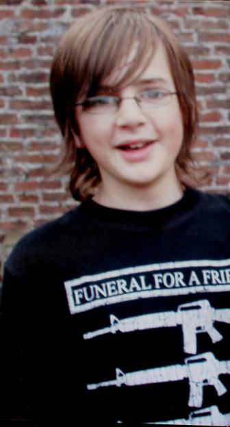 Andrew Gosden, a smiling young man with brown hair and glasses, wearing a black t-shirt with "FUNERAL FOR A FRIEND" printed on it.