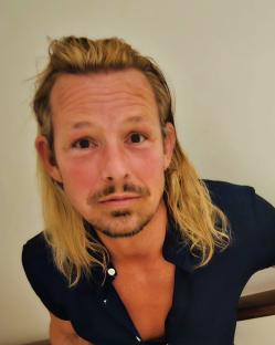 Headshot of Adam Rickitt with long blond hair.