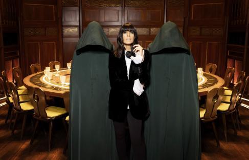 Claudia Winkleman standing between two figures in hooded cloaks at a round table.