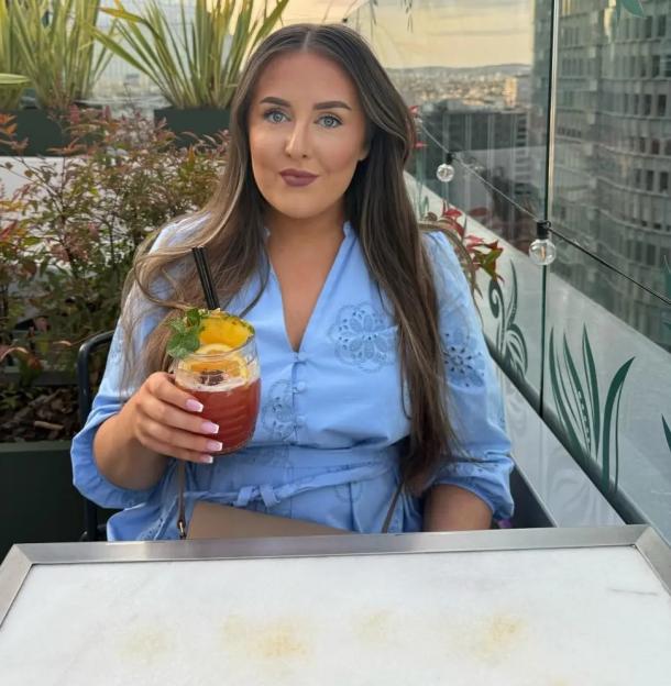A young woman in a blue dress with a drink, smiling at the camera on a rooftop bar.