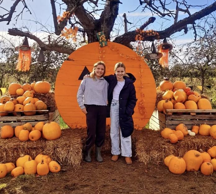Two girls pose in front of a giant pumpkin decoration and pumpkins at a pumpkin patch.