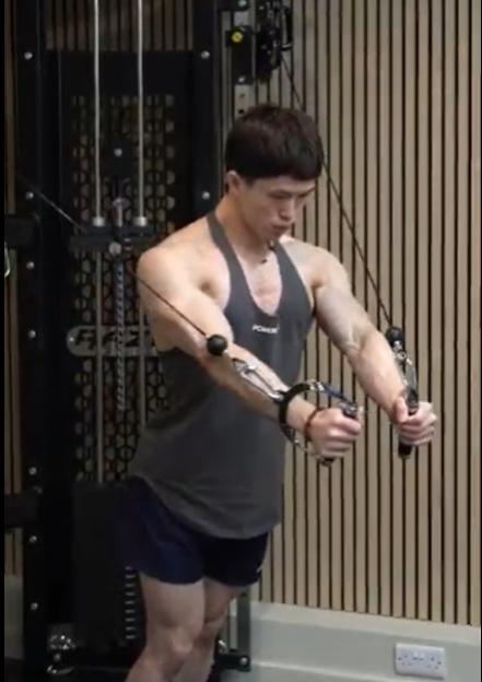 A man in a grey tank top and dark shorts performing a cable fly exercise at a gym.