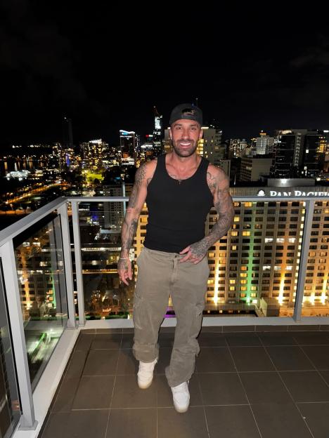 Billy Belcher standing on a balcony at night with a city skyline in the background.