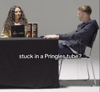 A man and woman sit at a table with Pringles cans, the man appearing to be attached to a chair by strings. The text