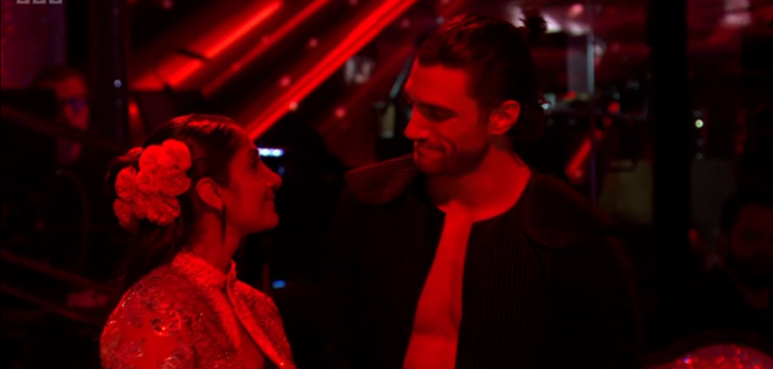 Two dancers in a dimly lit room, illuminated by red lights, looking at each other.