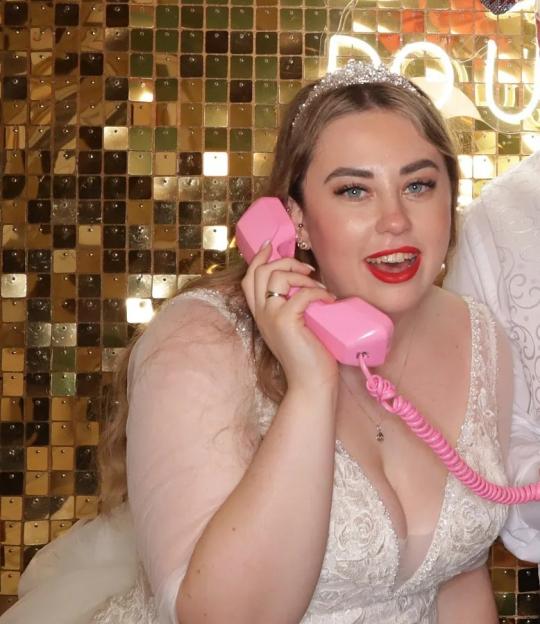 Bride and groom in wedding attire holding pink phones and a pineapple in front of a gold sequin backdrop.