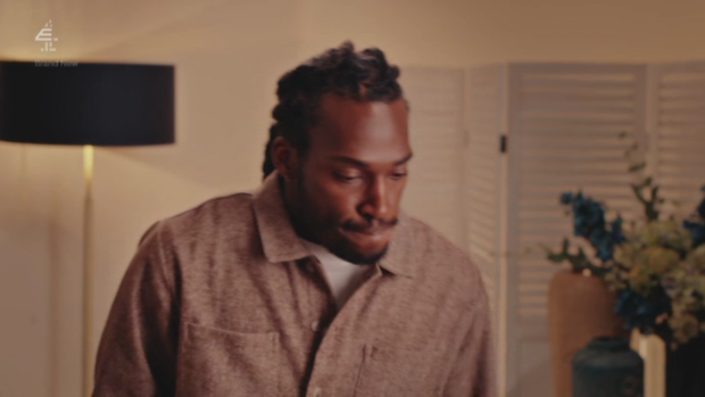 A man with dreadlocks looking down, in a room with a lamp and flowers.