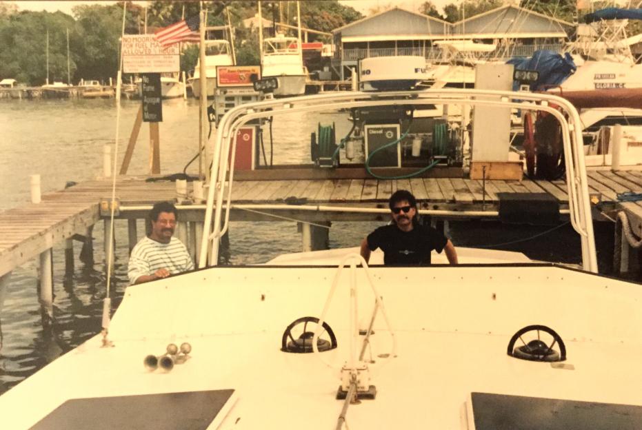 Two men on a boat at a dock with a fueling station.