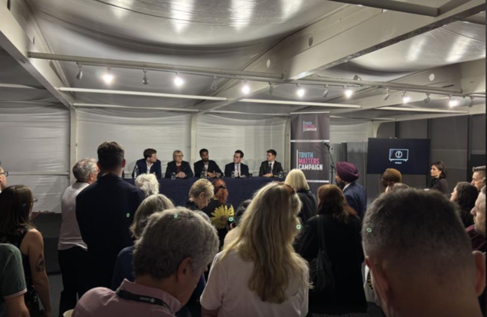 A panel of six people seated at a table in front of an audience, with a "Truth Matters Campaign" banner behind them.