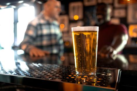 A close-up of a pint glass filled with lager. Unrecognisable people are sitting behind the drink conversing with each other. The lager is golden in colour creating foam and bubbles ready for the customer. Videos are available similar to this scenario