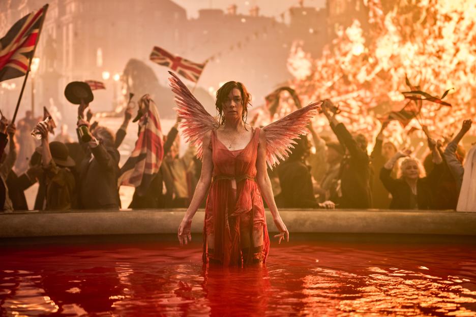 Woman in red dress with wings standing in a pool of red water, surrounded by a crowd.