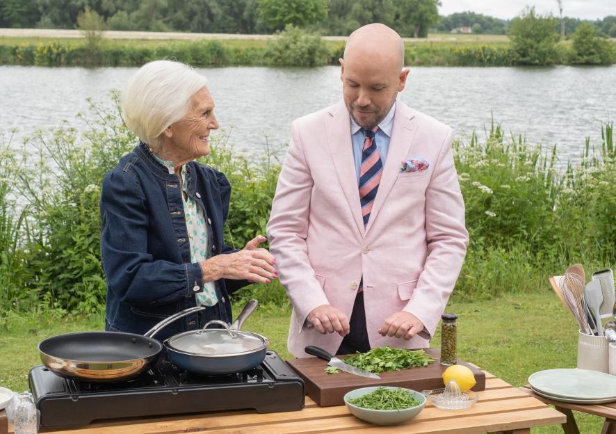 Mary Berry and a bald man in a pink suit cook outdoors beside a lake.