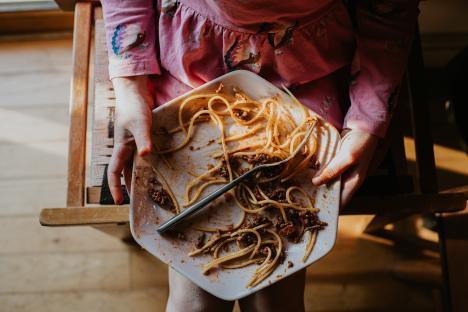 A plate of spaghetti bolognese rests on a child's knee. She has almost finished the meal, except for a few remaining strands of spaghetti. The image is taken looking directly down at the plate, with full focus being on the food and the fork.