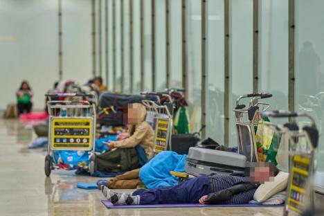 People sleeping on the floor of an airport terminal with luggage carts nearby.