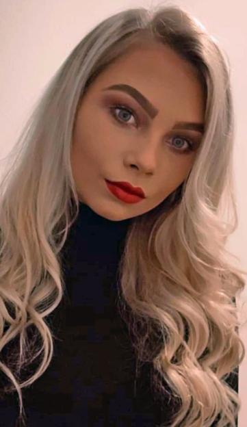 Close-up of a young woman with long blonde hair, red lipstick, and grey eyes, looking slightly to her left.