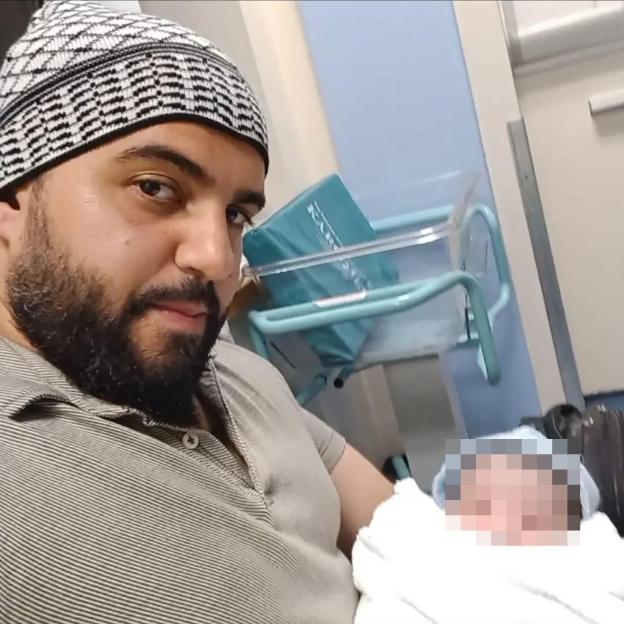 A man in a patterned cap holds a baby whose face is pixelated.