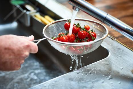 A hand rinsing vine tomatoes in a metal colander under running water.